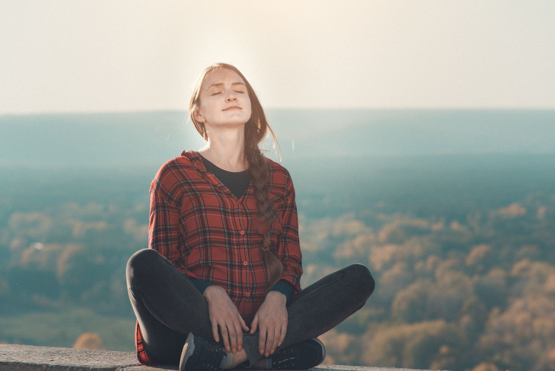 Jeune femme assise en tailleur, respirant l'air les yeux fermés. A l'arrière plan, se trouve un paysage automnal