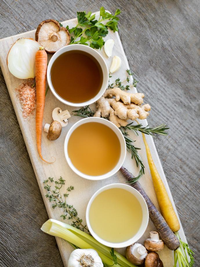 Trois bols posés sur une blanche en bois clair, comportant respectivement des liquides bruns, oranges et jaunes. Ils sont entourés de divers légumes (carottes, champignons, poireaux, ail, oignons et gingembre)