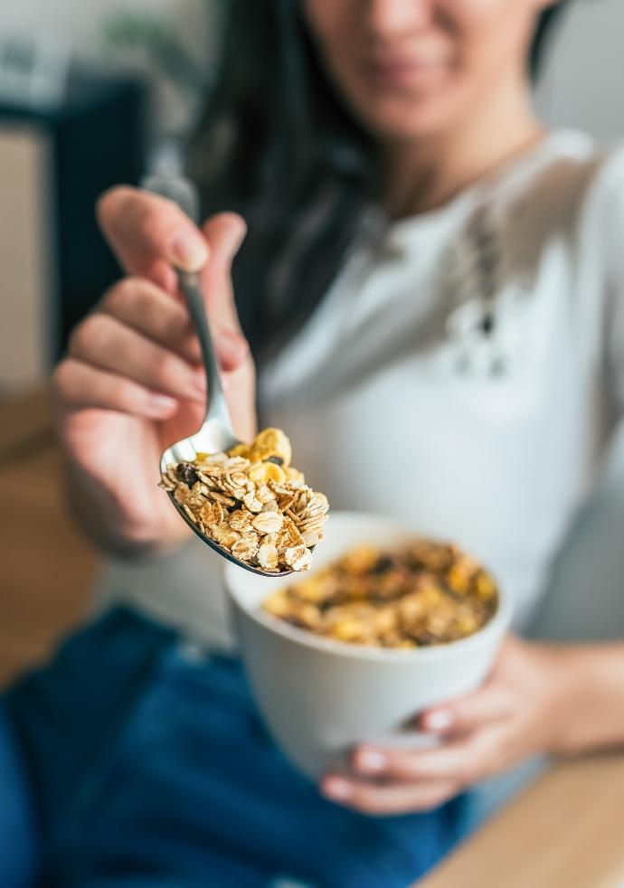 Jeune femme en arrière fond qui tient un bol et tend vers l'avant une cuillère de granola 